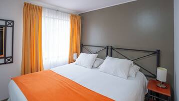 Una cama bien hecha con ropa de cama blanca y naranja, un cabecero de metal y una mesita de noche con una lámpara.