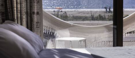 A hammock chair and a small table by a window overlooking a beach.