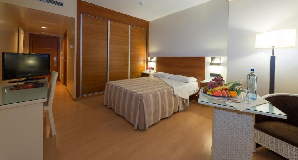 A hotel room with a bed, a desk with a lamp, a TV, and a fruit basket.
