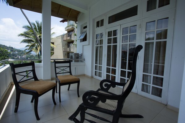 A balcony with two wooden chairs and a bench, white walls, and a view of a hill with buildings and palm trees. - Oak Ray Serene Garden Hotel (Kandy)