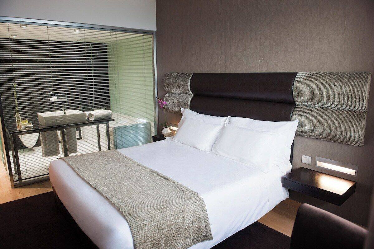 Ein modernes Hotelzimmer mit einem großen Bett, einem Nachttisch, einer Lampe, einem Fenster mit Jalousien und Blick nach außen.