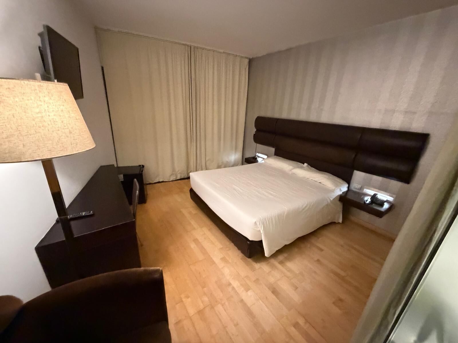 Habitación de hotel con cama, escritorio con lámpara, silla y ventana grande con cortinas.