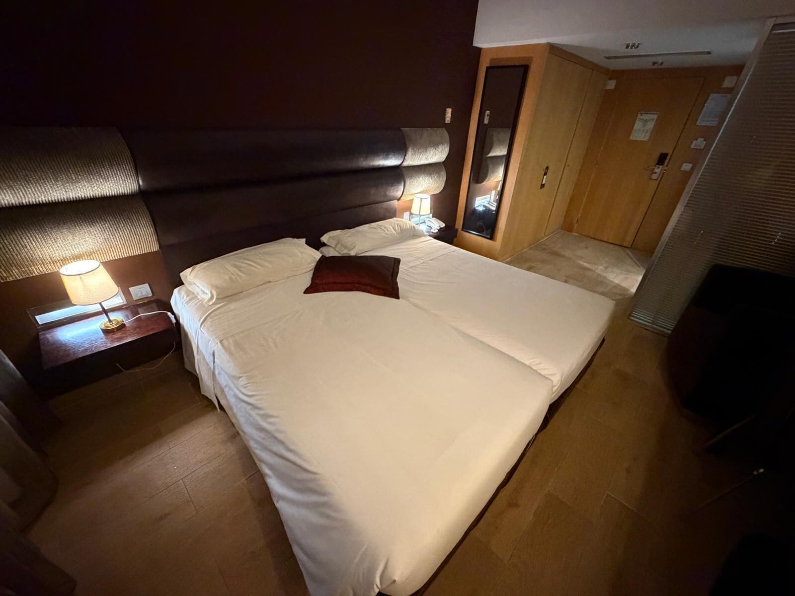 A hotel room with a large bed, a nightstand with a lamp, a wardrobe, and a mirror.
