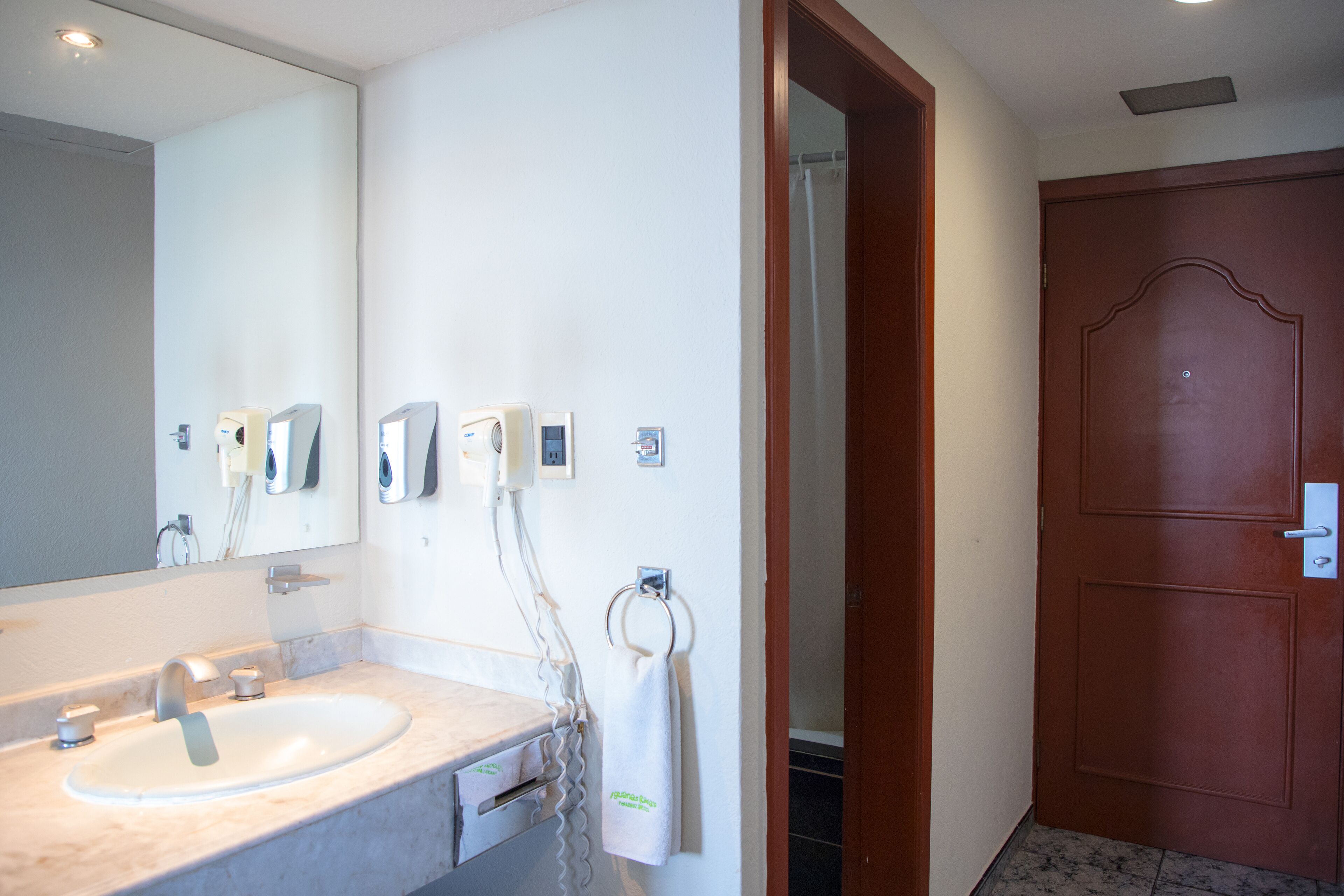 Un baño con lavamanos, espejo, dispensador de toallas y una puerta de madera.