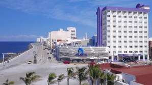 A coastal cityscape with a prominent white building with a purple roof, a large hotel with a blue logo, and a clear view of the ocean. - Hotel Lois Veracruz (Boca del Río)