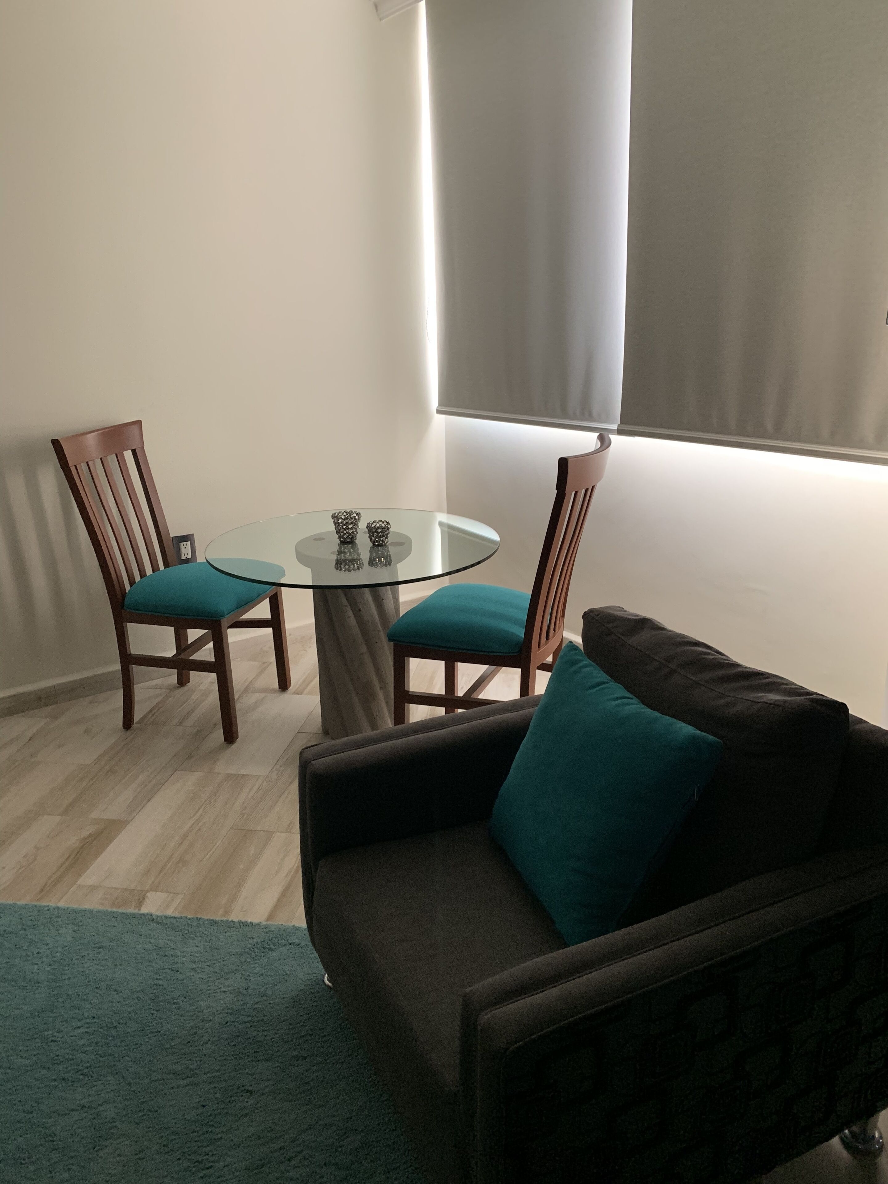 Una sala de estar con una mesa redonda de cristal, dos sillas de madera, un sofá oscuro con cojines verde azulado y una alfombra verde.