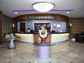 Zona de recepción de hotel con un cartel de 'Bienvenidos', un mostrador de recepción, un menú digital y una escultura decorativa en el centro.
