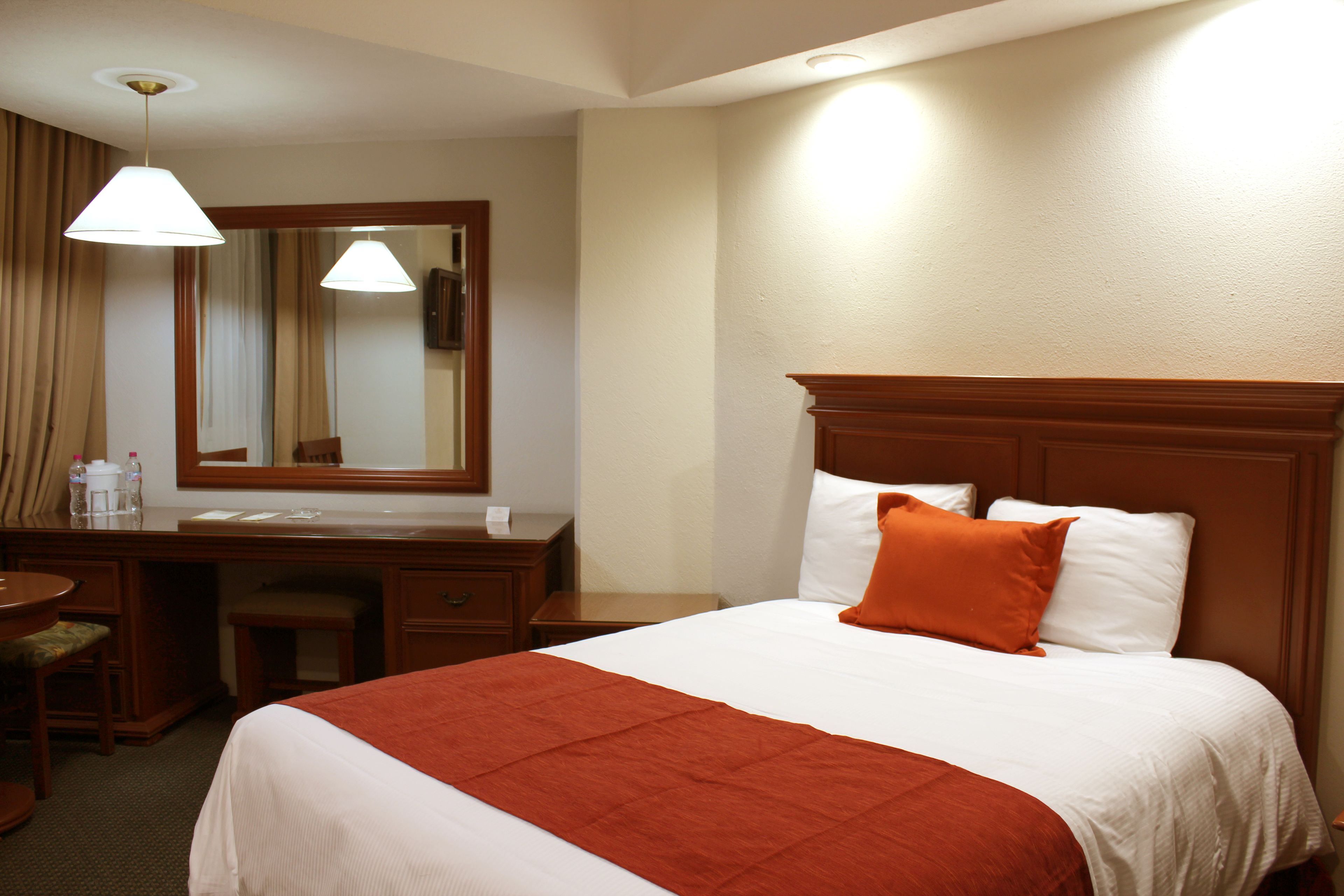 A hotel room with a bed, a desk, a chair, a mirror, and a lamp.
