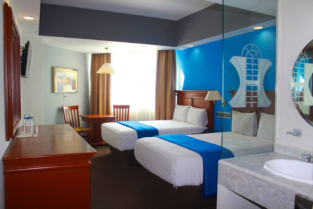 A hotel room with two beds, a desk, a chair, a lamp, a mirror, and a bathroom with a sink and mirror.