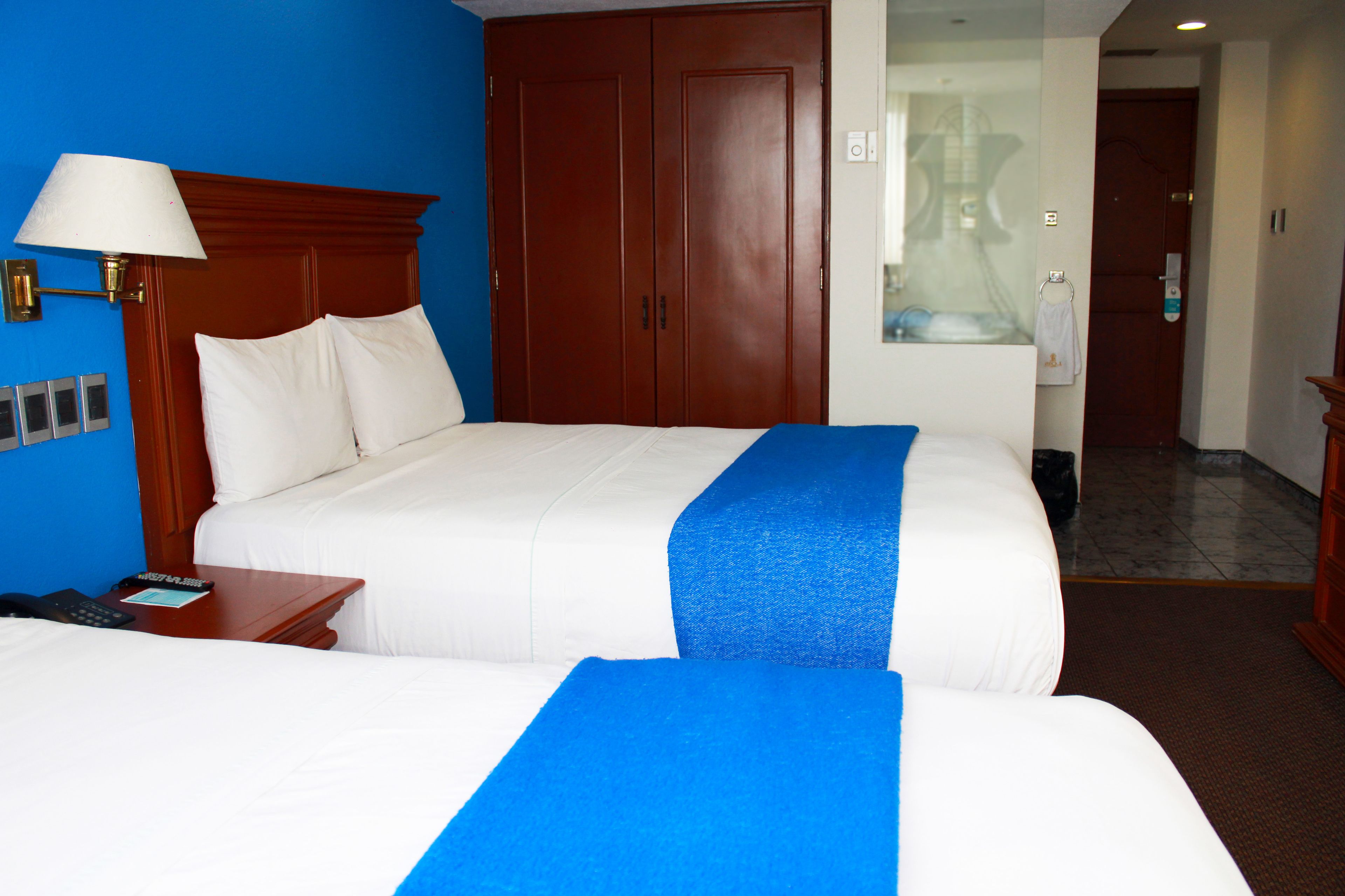 A hotel room with two beds, a wooden wardrobe, a bedside table with a lamp, and a bathroom visible through a glass door.