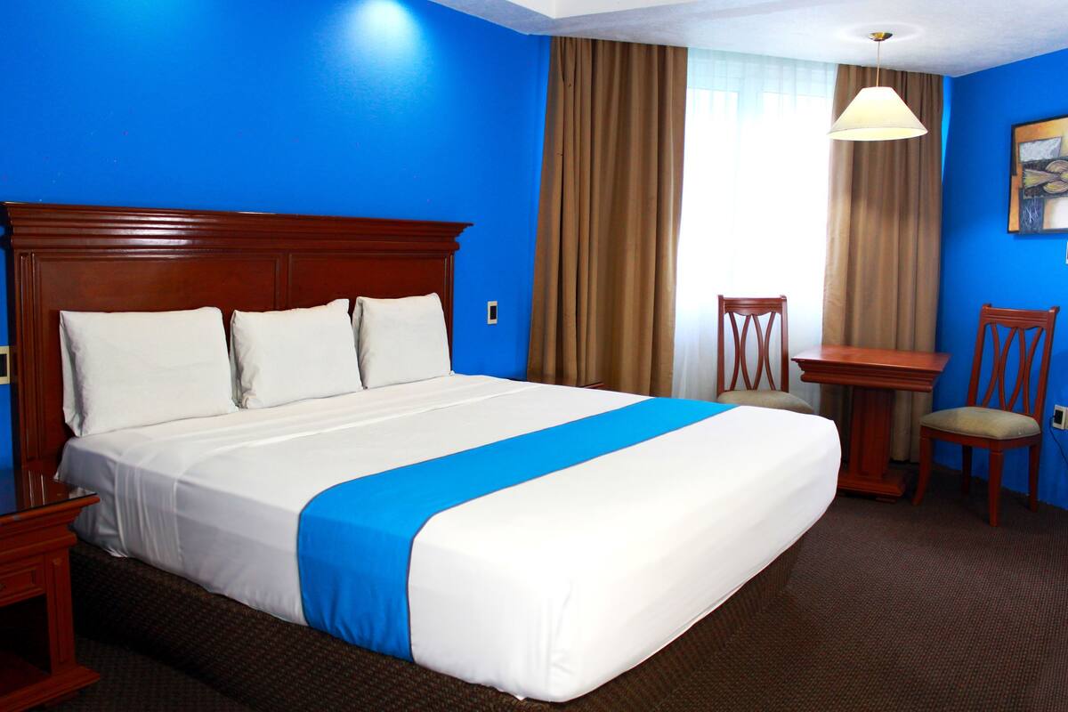 A hotel room with a large bed, a desk, two chairs, and a painting on the wall.