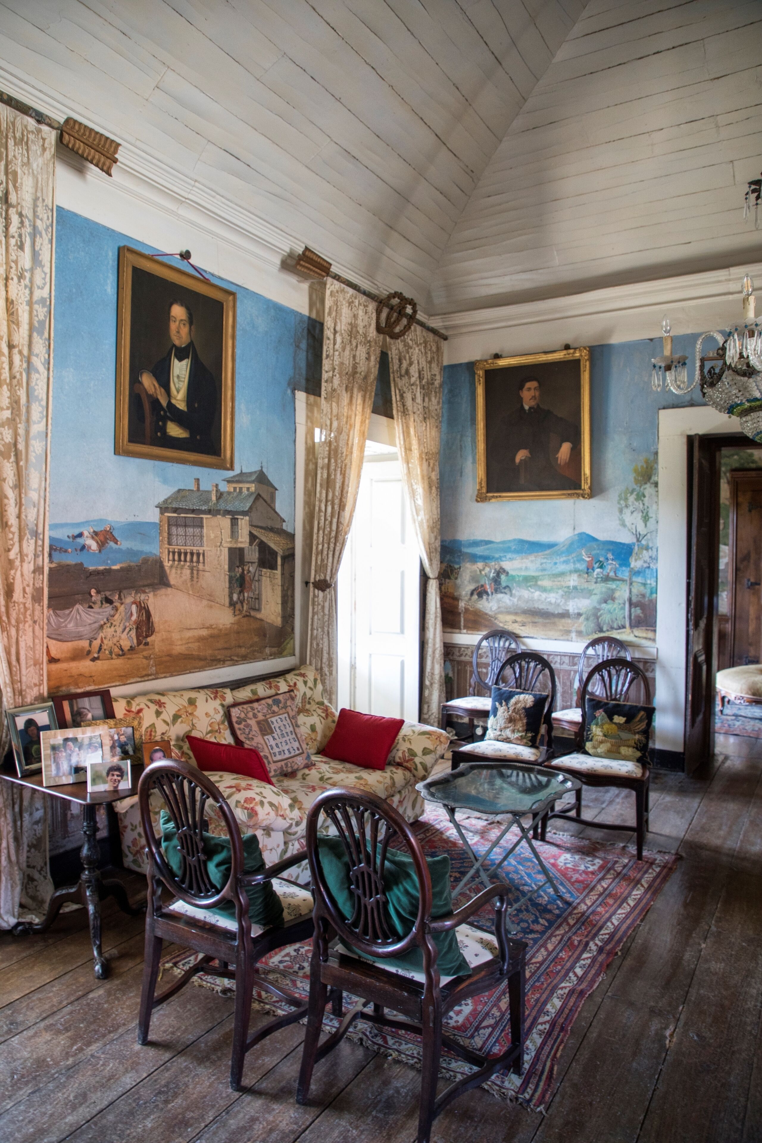 Una habitación con un cuadro grande de un paisaje, un sofá con tapicería floral y varias sillas de madera.