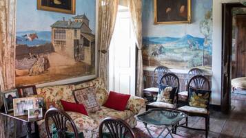 Una habitación con un cuadro grande de un paisaje, un sofá con tapicería floral y varias sillas de madera.