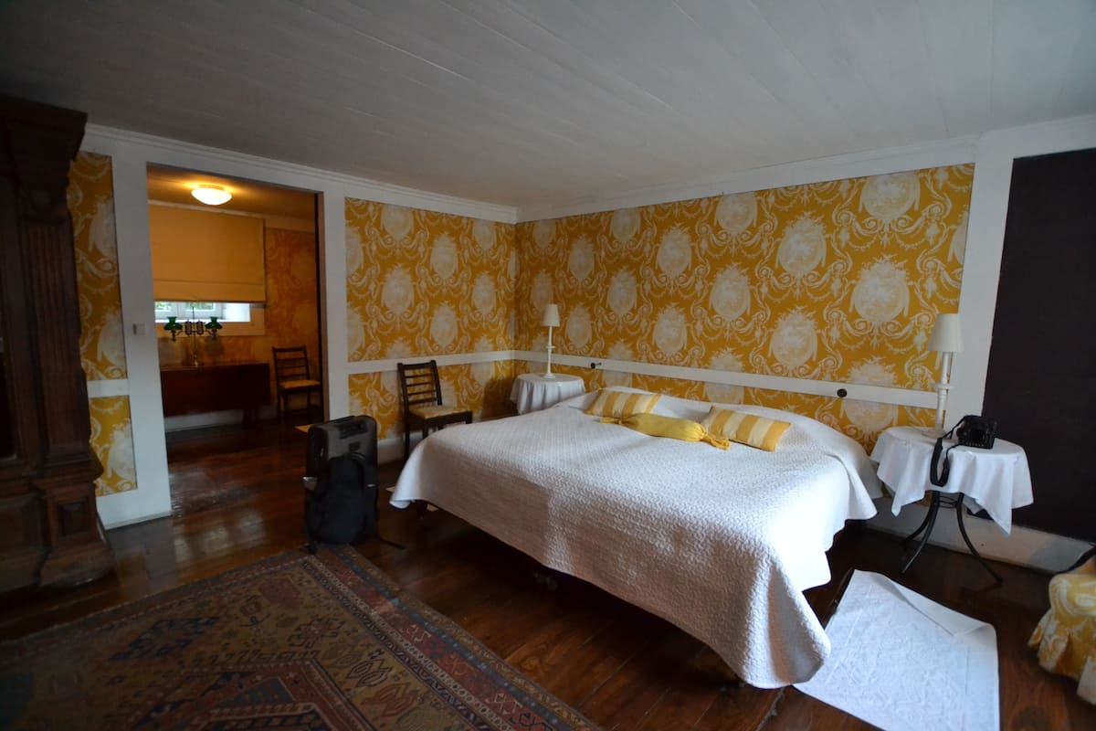 A hotel room with a large bed, a wooden floor, a patterned wall, and a small table with a lamp.