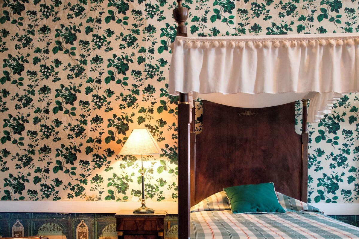 Una cama con dosel, una mesita de noche con lámpara, un papel tapiz estampado con motivos florales y un suelo de madera.