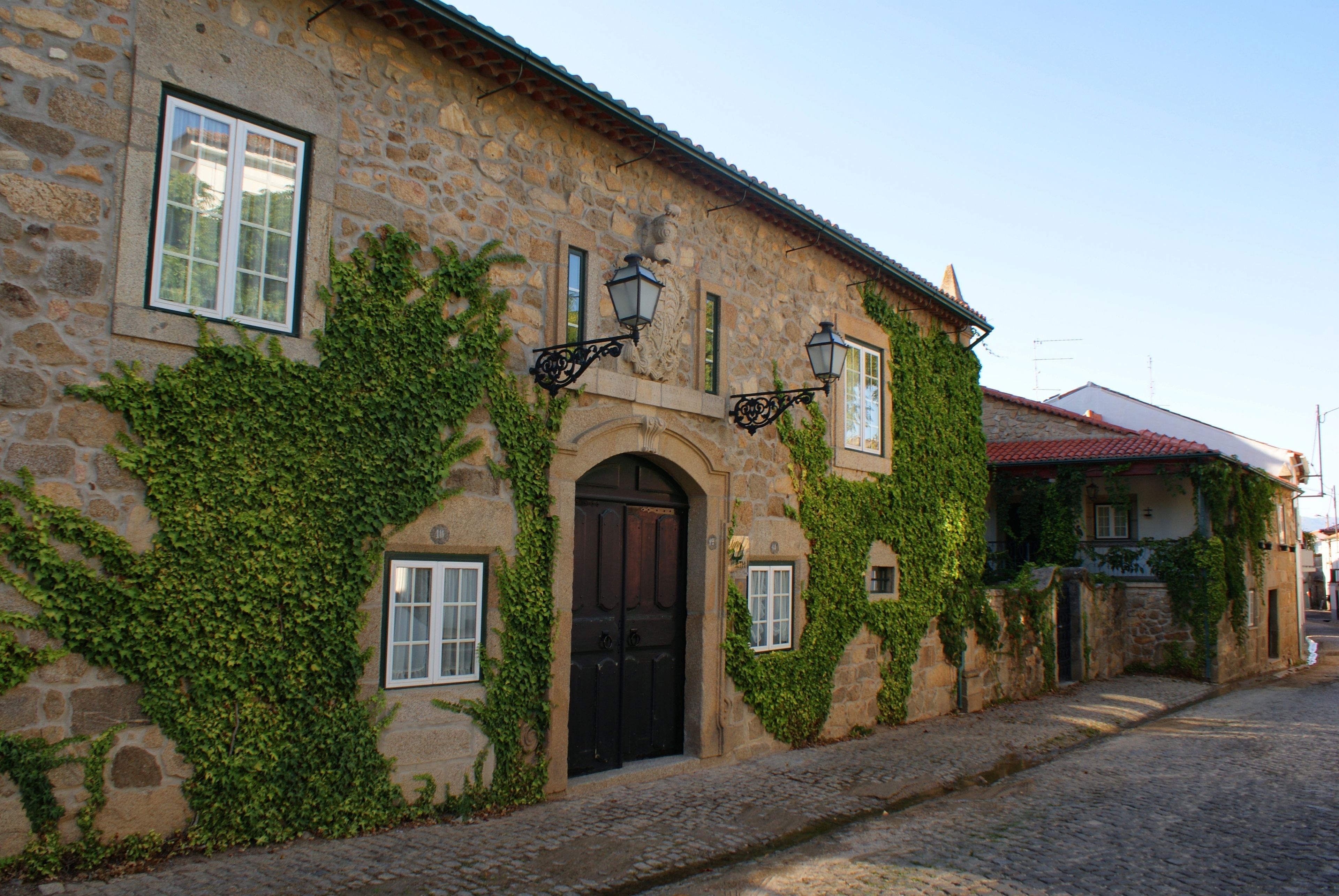Un edificio de piedra con muros cubiertos de hiedra, una calle adoquinada y una puerta negra tradicional.