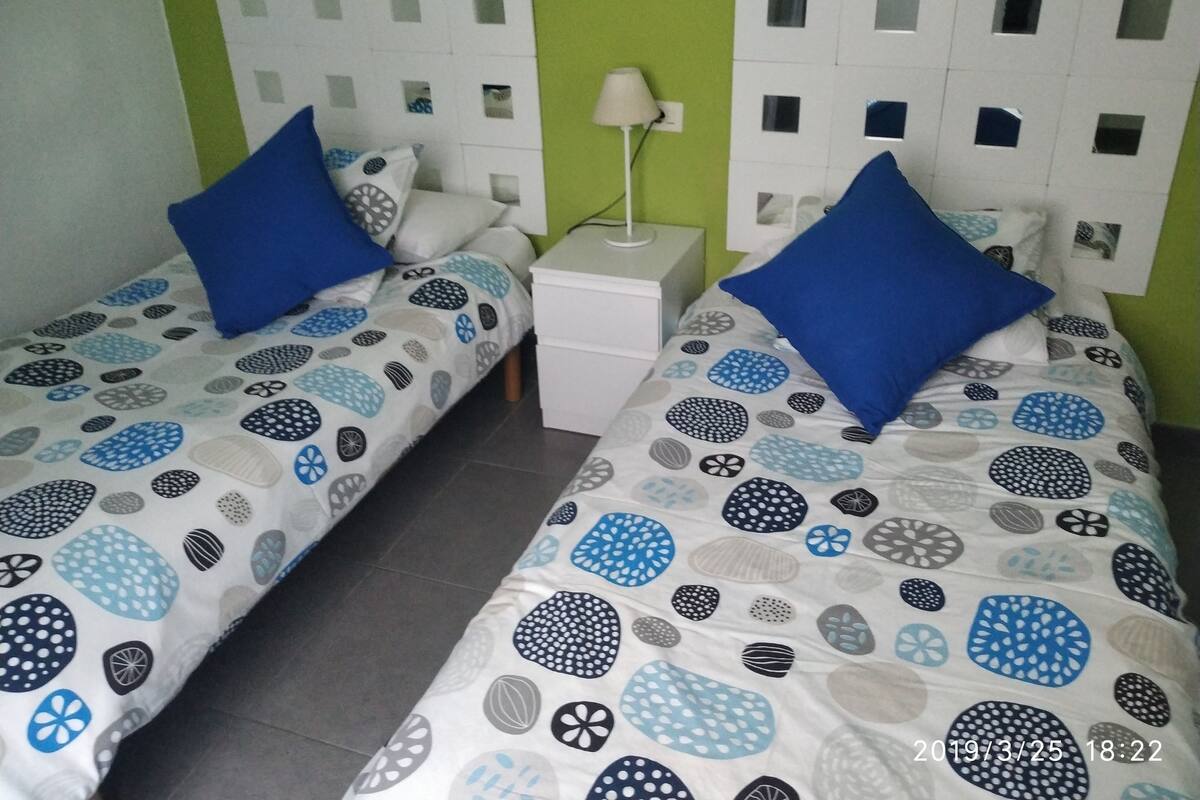A twin room with two single beds, each with a patterned bedspread and blue pillows. There is a white bedside table with a lamp between the beds.