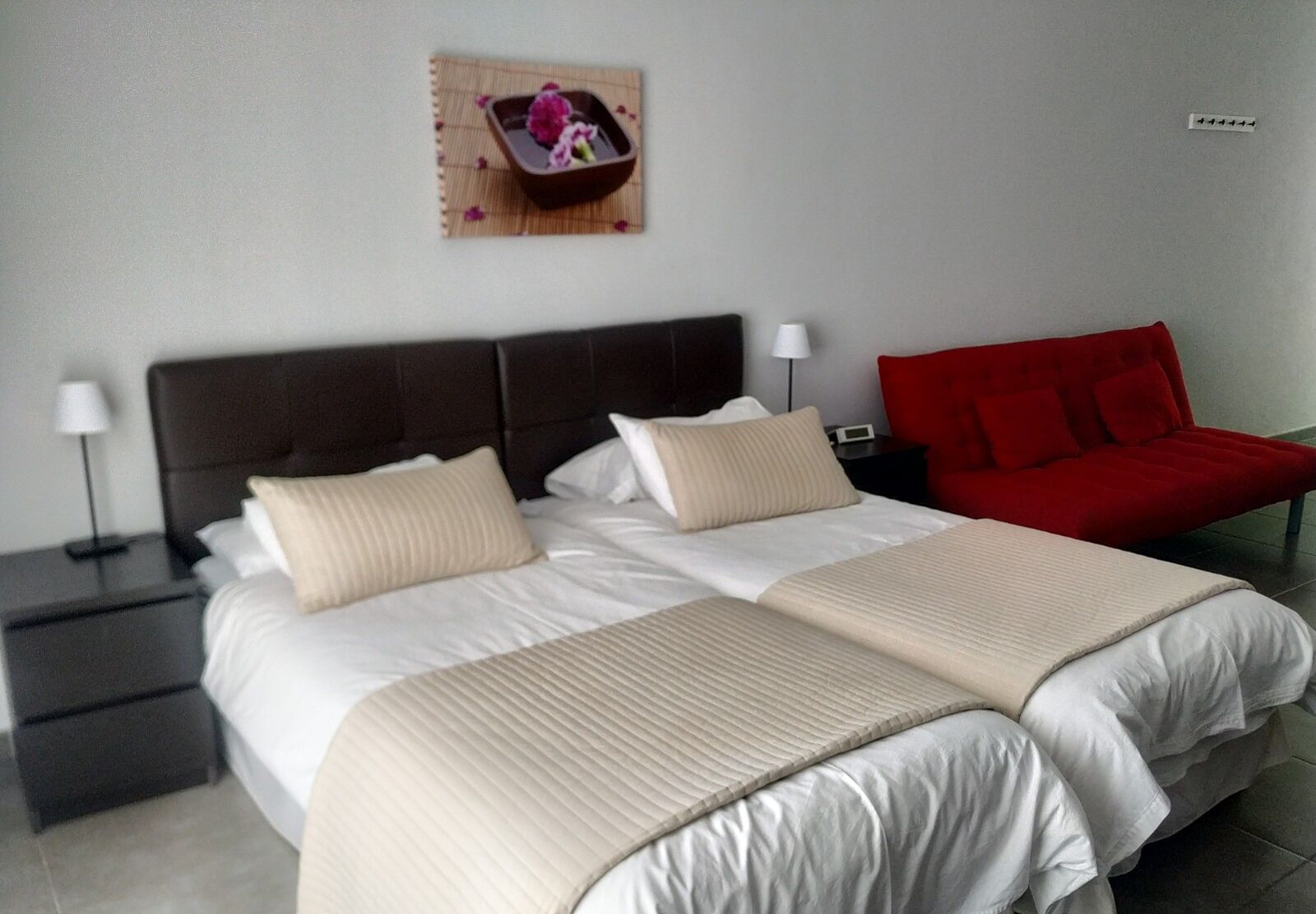 Una cama doble con dos almohadas, una mesita de noche con lámpara, una silla roja y una pequeña mesa con un arreglo floral.