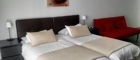 A double bed with two pillows, a bedside table with a lamp, a red chair, and a small table with a flower arrangement.