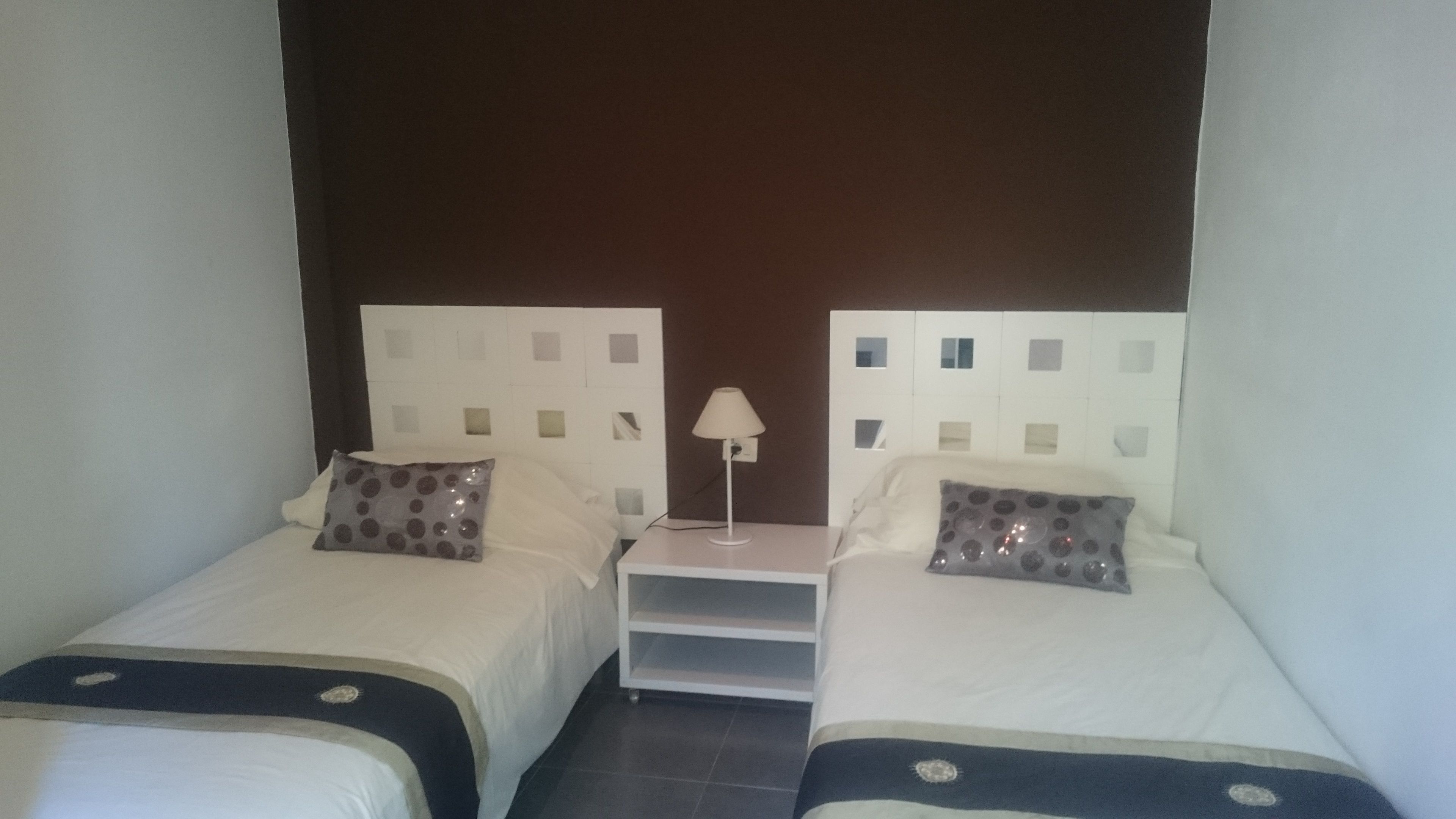 Een kamer met twee bedden, elk met een witte hoofdbord en een kussen met een rond patroon. Tussen de bedden staat een witte nachtkastje met een lamp erop.