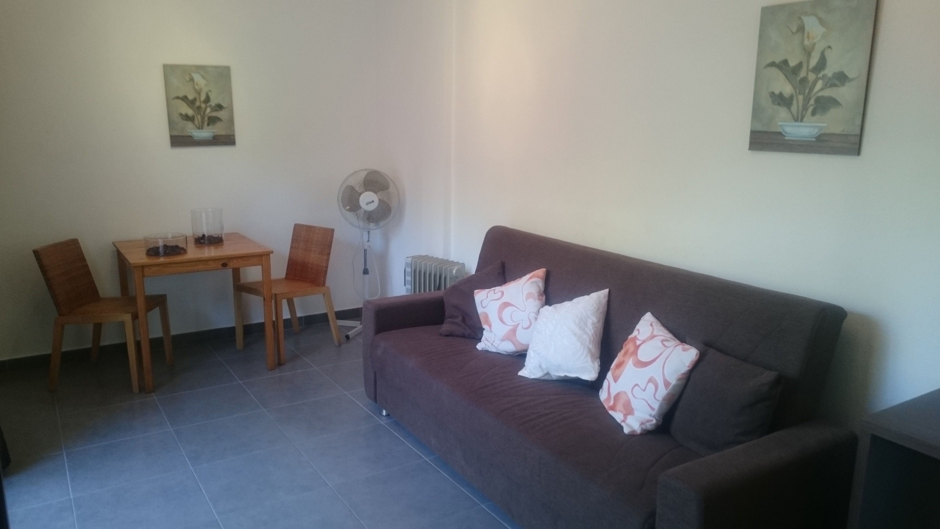 A living room with a brown sofa, two chairs, a small table, and a floor fan.