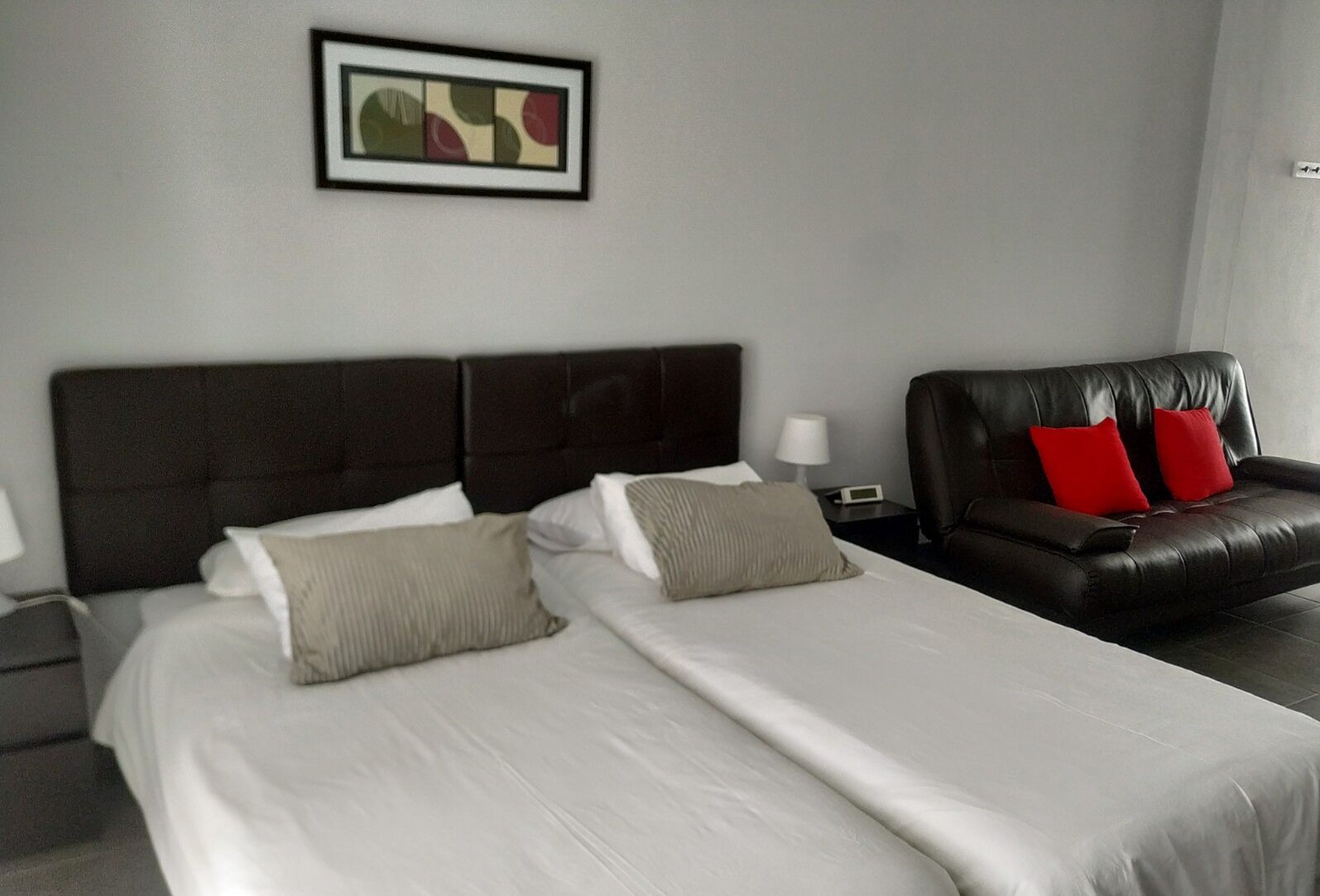 Een hotelkamer met een groot bed, een donkere leren bank met rode kussens, een nachtkastje met een lamp, en een lijst met een abstract schilderij aan de muur.