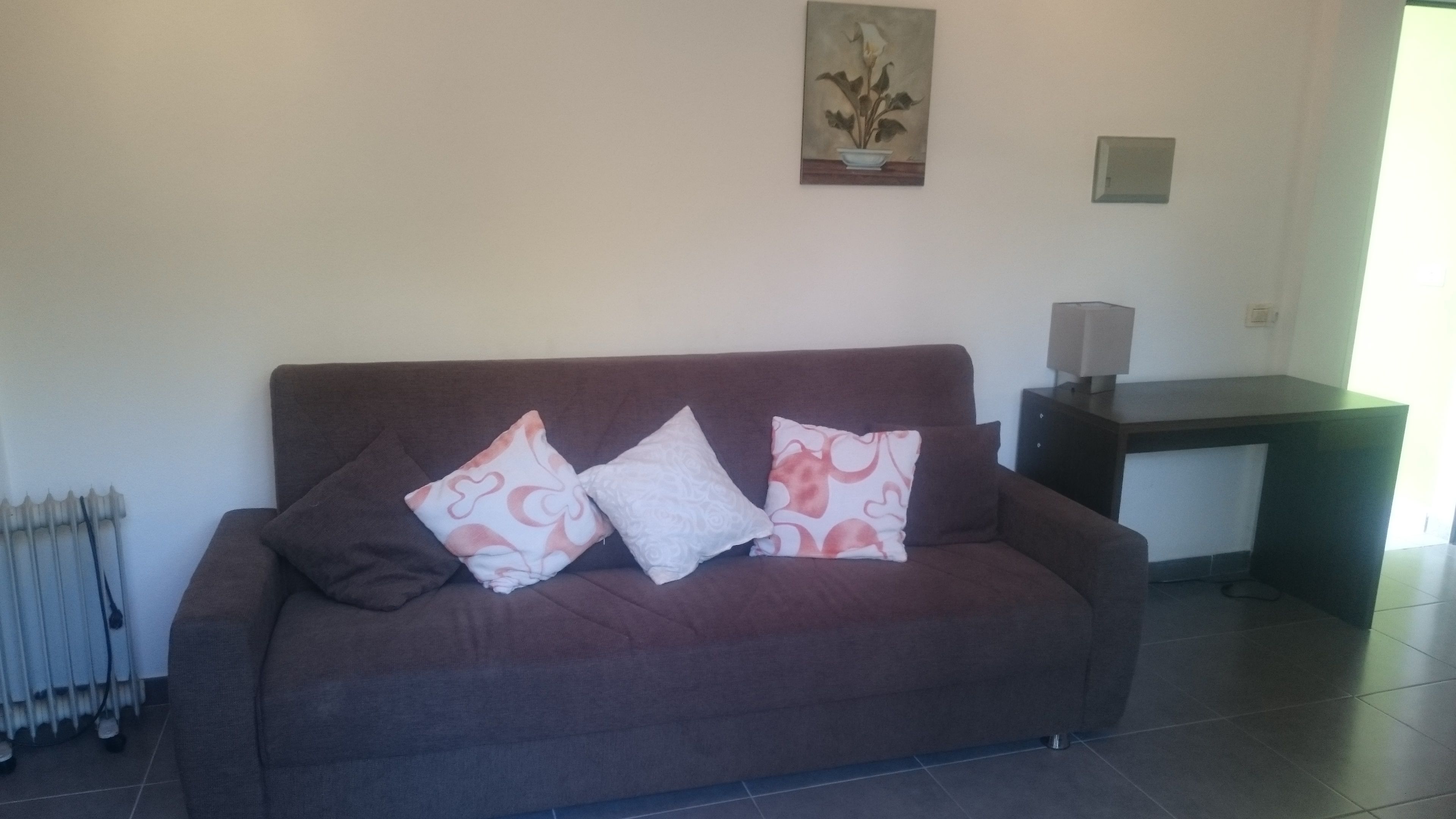 A brown sofa with three patterned cushions, a radiator, a painting, and a desk with a lamp.