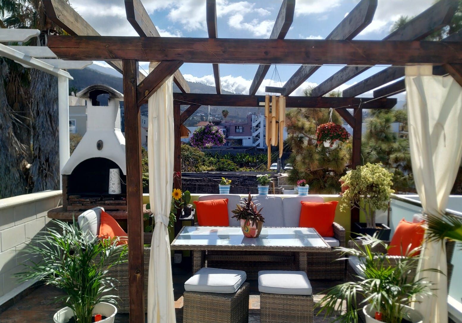 Un patio al aire libre con una pérgola de madera, un horno de pizza, muebles de mimbre y plantas en macetas.