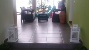 Un espacio al aire libre con techo, sillas y mesas, una pared verde y plantas en macetas.