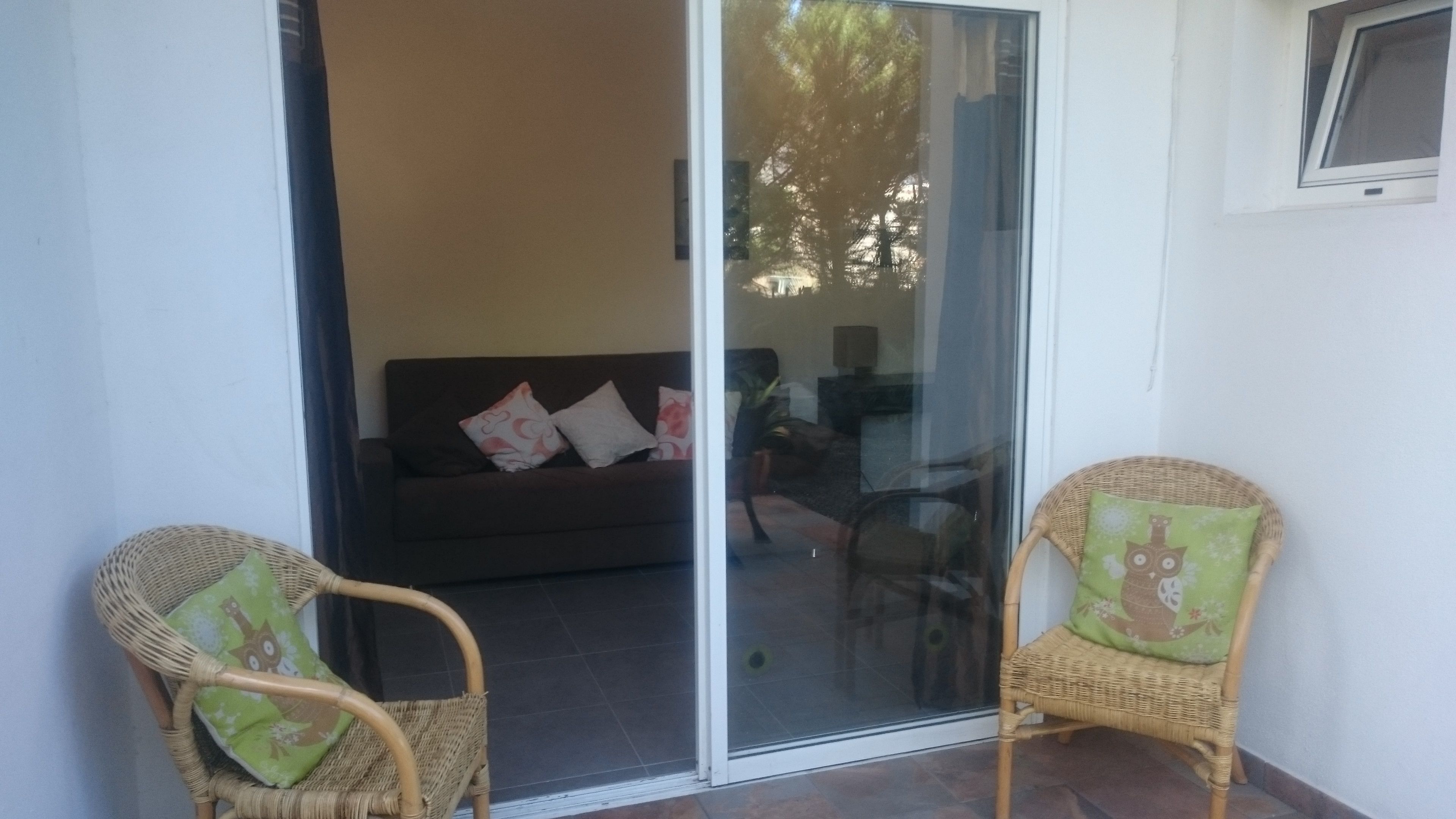 Una habitación con puerta corrediza de vidrio que da a un espacio exterior, un sofá con cojines y dos sillones de mimbre también con cojines.