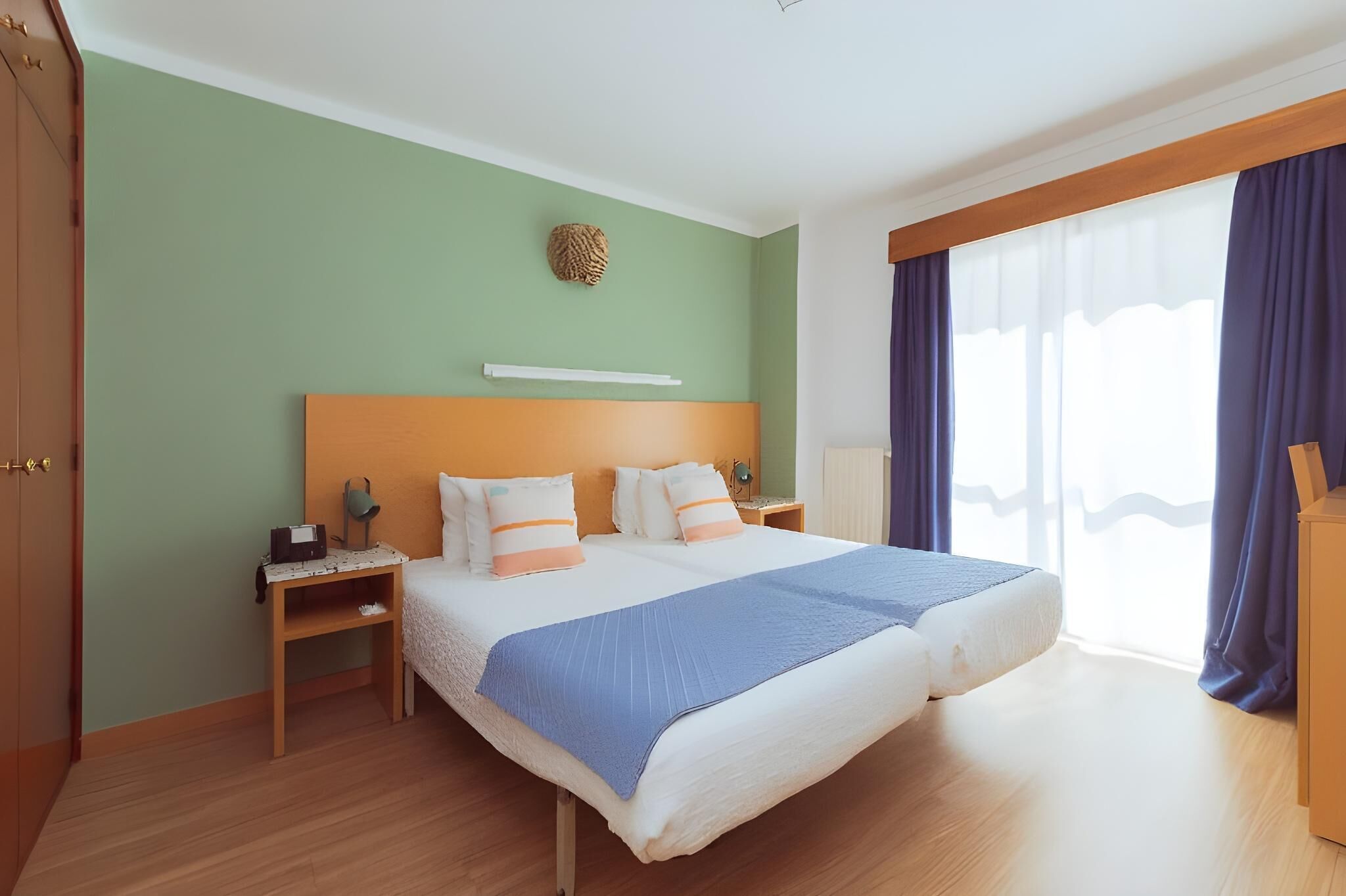 A double bed with a blue blanket, two bedside tables with lamps, a wooden headboard, a green wall, and a large window with curtains.