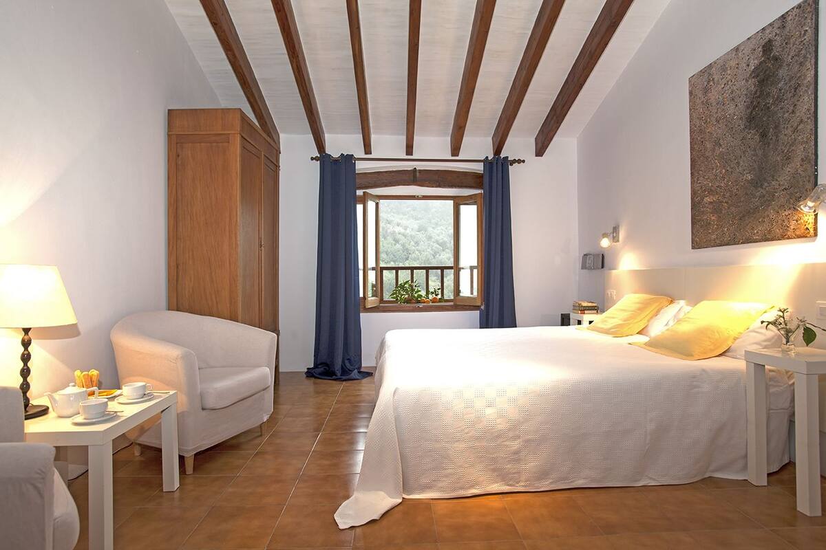 Une chambre à coucher avec un lit, une chaise, une table de chevet, une lampe et une fenêtre avec des rideaux.