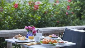 Een gedekte ontbijttafel met croissants, fruit, sap, een bloevas en een kop koffie op een balkon met uitzicht op groen en bloemen.