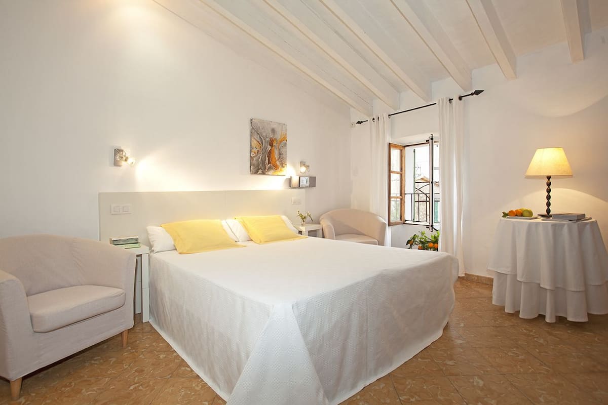 Une chambre d’hôtel avec un grand lit, une table de chevet, une chaise, une fenêtre avec des rideaux et une lampe sur une petite table.