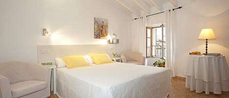 A hotel room with a large bed, a bedside table, a chair, a window with curtains, and a lamp on a side table.