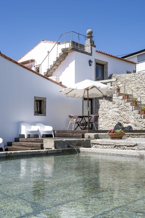 Zona de piscina con agua cristalina, sillas blancas, una mesa con sombrilla y una pared de piedra con escaleras.