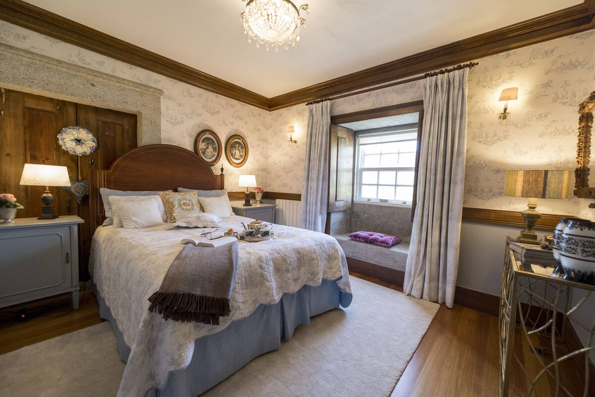 Une chambre à coucher avec un grand lit, une fenêtre avec des rideaux, un lustre et une table de chevet avec une lampe.