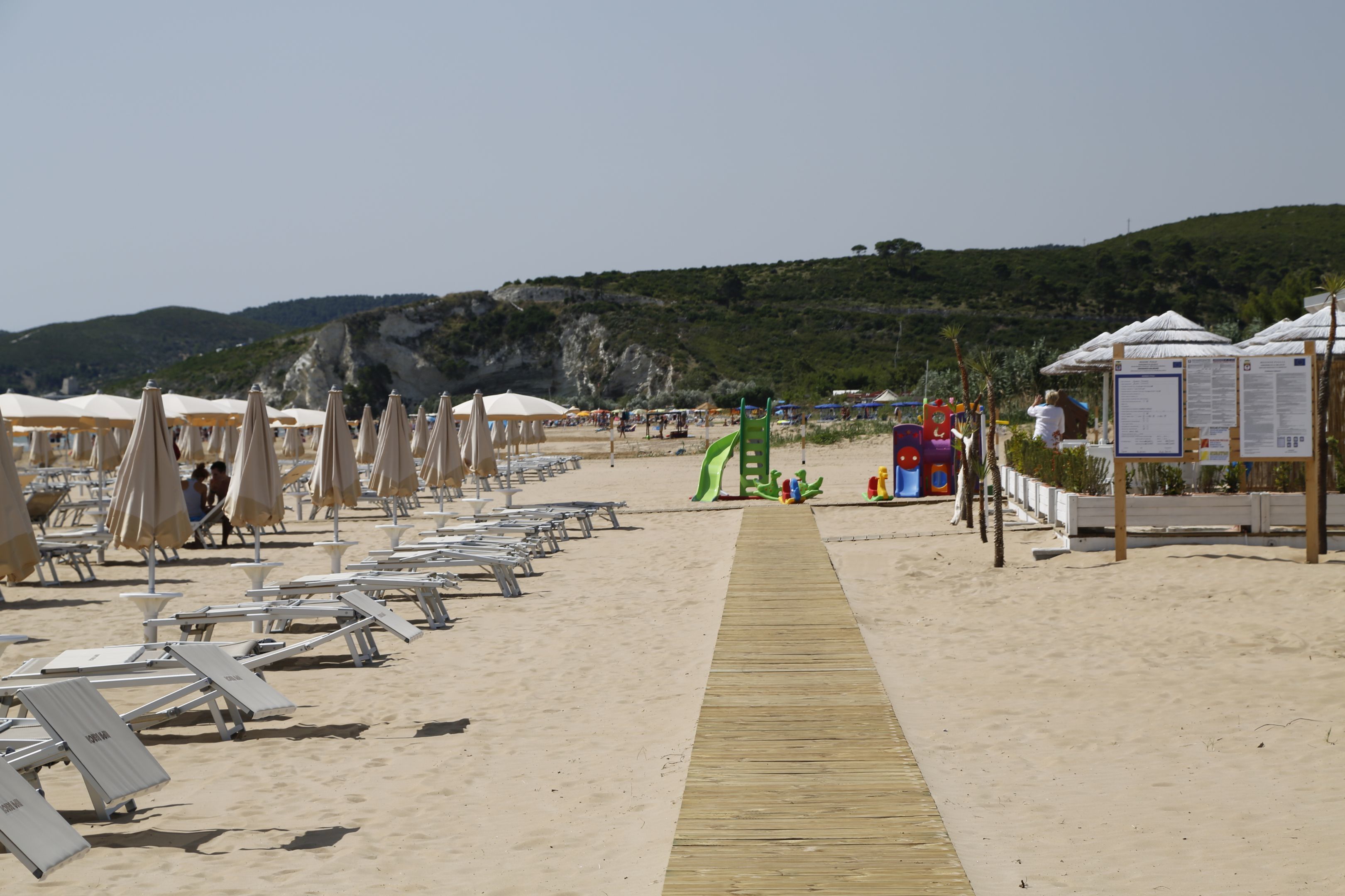 Una playa con filas de tumbonas y sombrillas, un pasillo de madera y un parque infantil.
