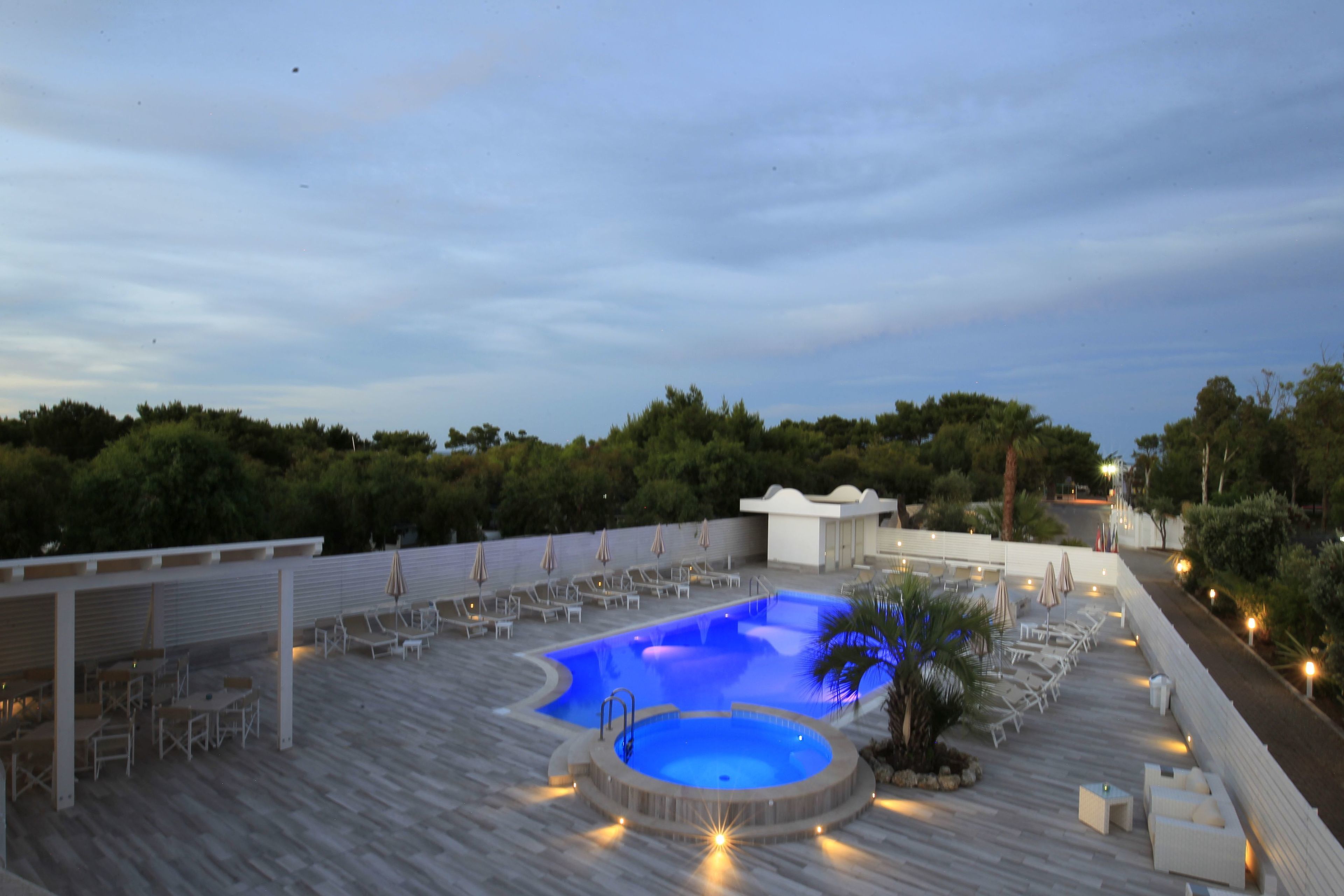 Un área de piscina en la azotea con sillones de descanso y sombrillas, rodeada por una terraza de madera y una pared blanca.