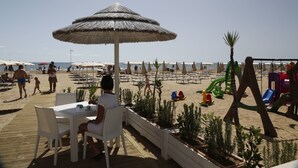 En restaurang vid stranden med uteservering, ett parasoll med halmtak och en lekplats med rutschkanor.