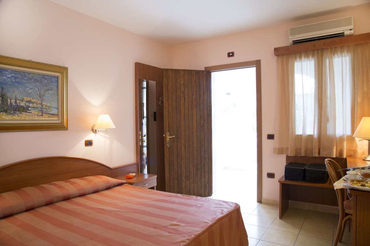 Une chambre d’hôtel avec un lit, un bureau et une chaise, un tableau au mur et une porte donnant sur l’extérieur.