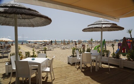 An outdoor seating area with white tables and chairs, covered by large thatched umbrellas, overlooking a sandy beach with umbrellas and lounge chairs.