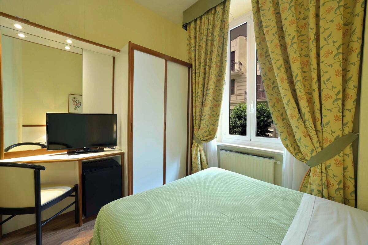 Een hotelkamer met een bed, een bureau met televisie, een stoel en een raam met gordijnen.