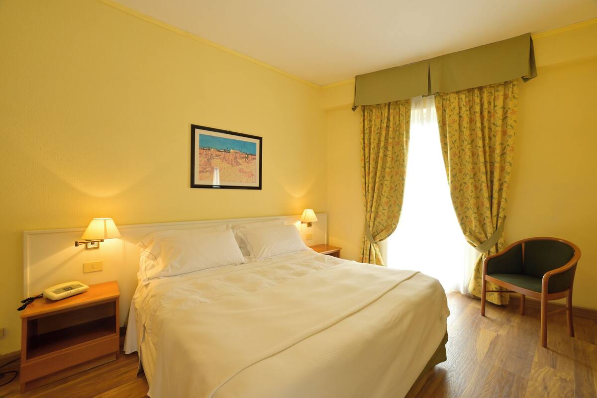 Een hotelkamer met een bed, twee bedlampen, een stoel, een nachtkastje met een telefoon en een lijst met een foto aan de muur.