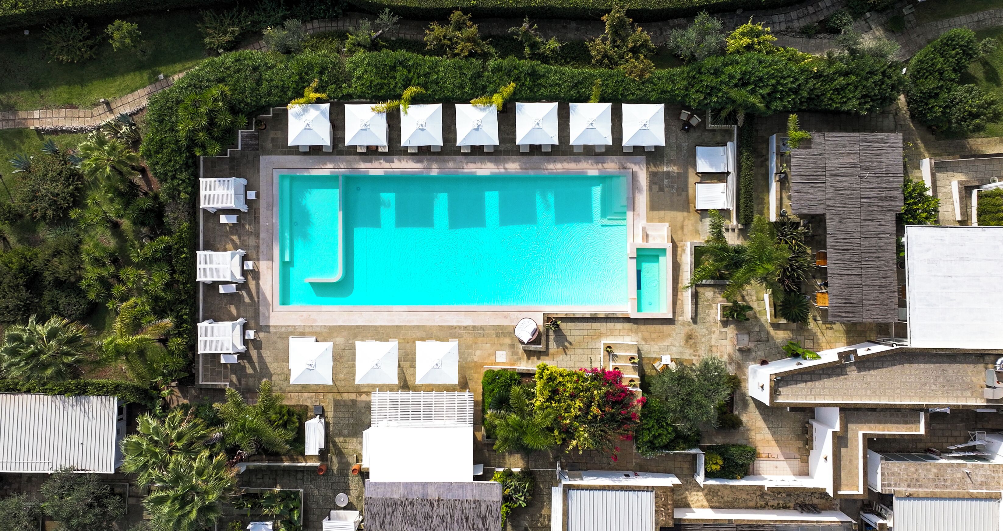 An aerial view of a swimming pool surrounded by lounge chairs and umbrellas, with a stone wall and greenery.