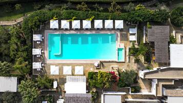 An aerial view of a swimming pool surrounded by lounge chairs and umbrellas, with a stone wall and greenery.