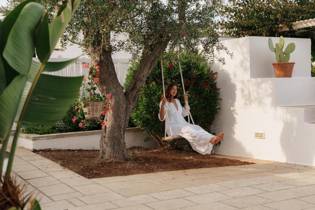 Une personne est assise sur une balançoire dans un jardin, avec un mur blanc et un pot de fleurs à proximité.