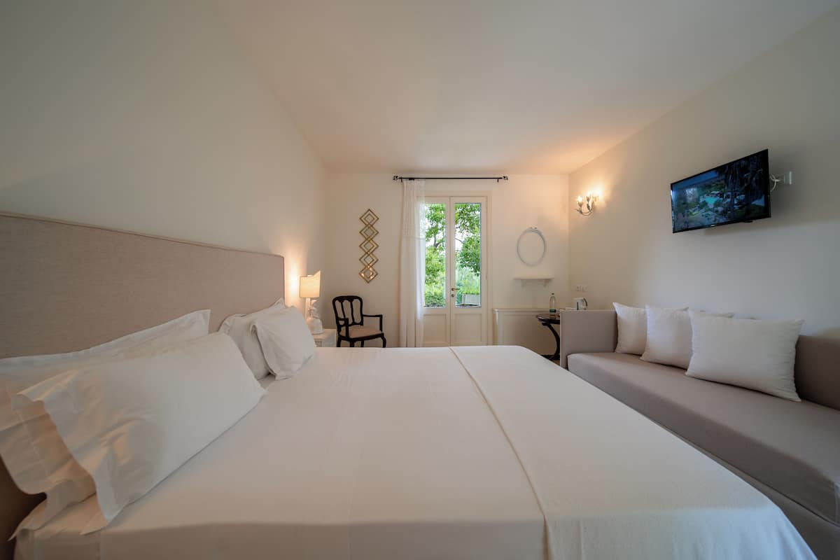 A hotel room with a large bed, a sofa, a desk, and a chair. There is a window with curtains, a painting on the wall, and a lamp on the desk.