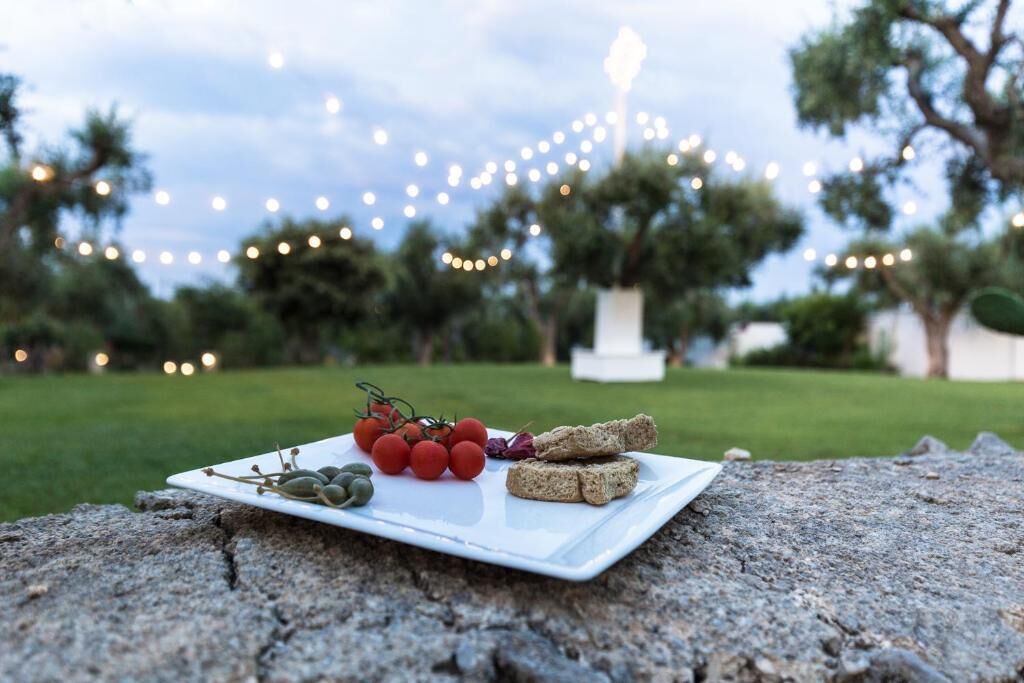 A white plate with tomatoes, olives, and bread on a rock outdoors with string lights in the background.
