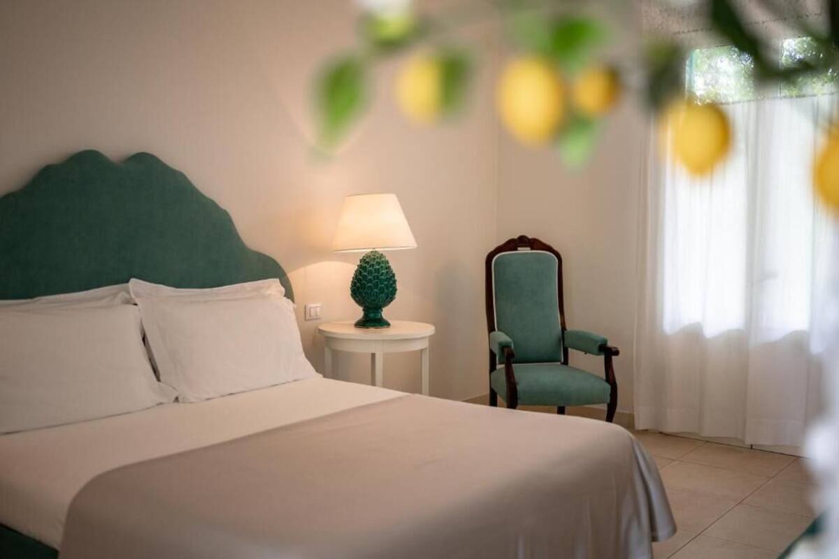 A neatly made bed with white linens, a green upholstered headboard, a small round table with a green lamp, and a teal chair.